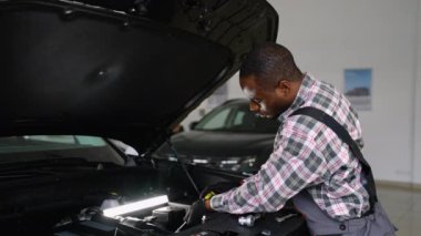 Tulumlu ve eldivenli Afrikalı bir oto tamircisi modern bir atölyede parlak bir iş lambası altında düzgün bir teşhis ve bakım sağlayan bir araba motoru tamir ediyor..