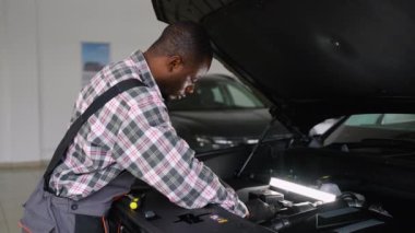 Tulumlu ve eldivenli Afrikalı bir oto tamircisi garajdaki bir iş lambasının altındaki araba motorunu tamir ediyor. Teşhis, bakım ve sorun giderme araçları kullanıyor..