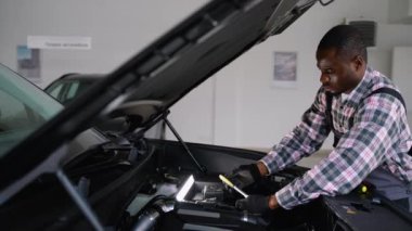 Tulum ve eldiven giyen Afrikalı bir oto tamircisi modern bir atölyede araç gereç ve çalışma lambası kullanarak bir araba motorunu onarıyor..