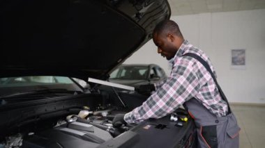 Eldiven ve tulum giyen profesyonel bir oto tamircisi modern bir atölyede çalışma lambası kullanarak bir araba motorunu inceler ve tanılama ve onarımlara odaklanır..