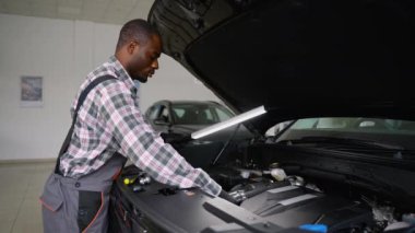 Eldiven ve tulum giyen profesyonel bir oto tamircisi modern bir atölyede parlak bir LED çalışma lambası kullanarak bir araba motorunu inceler ve düzgün bir teşhis ve bakım sağlar..