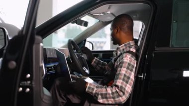 Eldiven ve tulum giyen profesyonel bir oto tamircisi modern bir atölyede teşhis ve sistem analizi için dizüstü bilgisayar kullanarak arabanın içinde oturur..