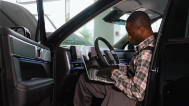 Profesyonel bir atölyede teşhis ve sistem analizi için bir laptop kullanan, tulumlu ve eldivenli bir Afrikalı Amerikalı otomobil tamircisi modern bir arabanın içinde oturur..