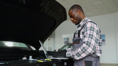 Tulum ve eldiven giyen Afrikalı bir oto tamircisi, profesyonel bir atölyedeki araba motoruna teşhis koymak için dizüstü bilgisayarda çalışır ve verimli bir araç bakımı sağlar..