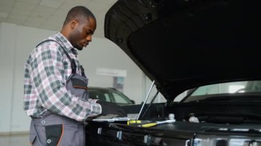 Tulum ve eldiven giyen Afrikalı bir oto tamircisi, profesyonel bir atölyedeki araba motoruna teşhis koymak için dizüstü bilgisayarda çalışır ve verimli bir araç bakımı sağlar..