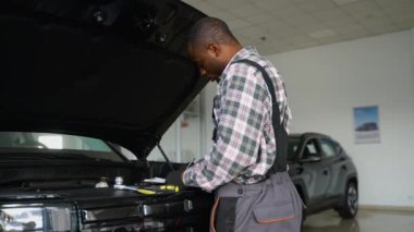 Tulum ve eldiven giyen profesyonel bir oto tamircisi, modern bir atölyede bir araba motorunu incelerken not alır..