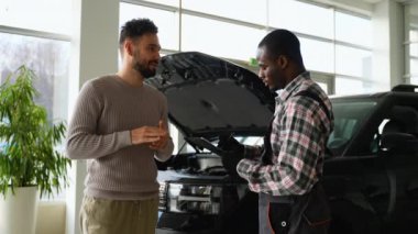 Tulum giymiş bir oto tamircisi, arka planda bir araç bulunan modern bir atölyede araba tamiri hakkında müşteriyle konuşur..