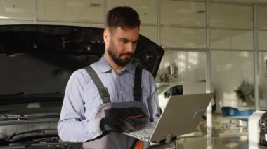 Servis merkezindeki profesyonel bir oto tamircisi, kaputu açık bir arabanın bakımını sağlamak için dizüstü bilgisayar kullanır..