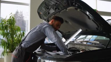 Otomobil tamircisi, profesyonel bir hizmet merkezinde detaylı teftiş için parlak bir LED çalışma ışığı kullanarak açık kaput altında bir araba motorunu inceler..