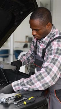 Tulum ve eldiven giyen Afrikalı bir oto tamircisi modern bir atölyede tamire başlamadan önce çalışma lambasıyla bir araba motorunu inceler..