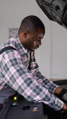 Tulum ve eldiven giyen Afrikalı bir oto tamircisi modern bir atölyede araç gereç ve çalışma lambası kullanarak bir araba motorunu onarıyor..