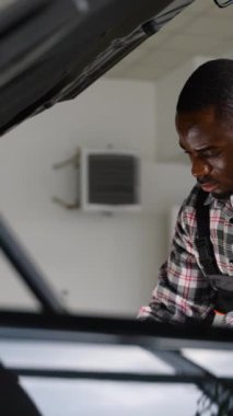 Eldiven ve tulum giyen profesyonel bir oto tamircisi, modern bir atölyede motor bakımı ve onarımı yapan bir arabanın kaputu altında konsantrasyonla çalışır..