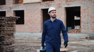 Başlık takan ve mavi üniformalı bir erkek inşaat işçisi bitmemiş bir binayla aktif bir şantiyede güvenle yürüyor..