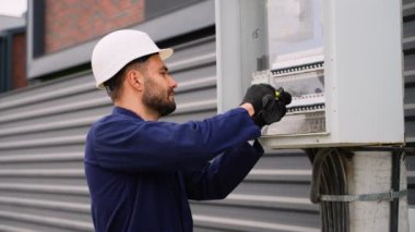 Beyaz bir şapka ve eldiven takan profesyonel bir elektrikçi bir inşaat alanındaki açık hava elektrik panelinin içine kablo döşeyip ayarlar..