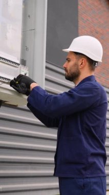 Beyaz bir şapka ve eldiven takan profesyonel bir elektrikçi modern bir çift katlı kulübe yakınına bir açık hava elektrik paneli kurup ayarlıyor.