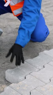 Mavi tulumlu ve güvenlik teçhizatlı bir inşaat işçisi, plastik bir çekiç ve ruh seviyesi kullanarak kumlu bir yüzeye birbirine kenetlenmiş taşlar yerleştirir..
