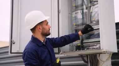 Sert şapka ve eldiven takan profesyonel bir elektrikçi, ayar için bir tornavida kullanarak açık hava elektrik panelini kontrol edip onarıyor..