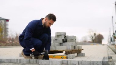 Donanma üniformalı bir inşaat işçisi kaldırıma kaldırım taşları yerleştiriyor, kauçuk bir çekiç kullanıyor ve düzgün hizalamak için düzeyi ayarlıyor..