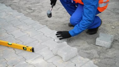 Mavi tulumlu ve güvenlik teçhizatlı bir inşaat işçisi, plastik bir çekiç ve ruh seviyesi kullanarak kumlu bir yüzeye birbirine kenetlenmiş taşlar yerleştirir..