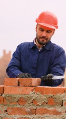 Kırmızı şapkalı ve mavi üniformalı bir inşaat işçisi, arka planda tamamlanmamış evler bulunan bir inşaat sahasında malayla tuğla örüyor..
