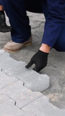 Mavi tulumlu bir inşaat işçisi bir yola kenetlenmiş kaldırım taşları serer. Bir çekiç ve sarı seviye bir alet hassasiyet için kullanılır..