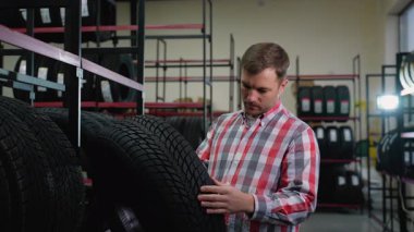 Ekose gömlekli bir adam otomobil parçası dükkanından almadan önce lastiği dikkatle inceler. Etrafı çeşitli lastik seçenekleriyle dolu raflarla çevrilidir..