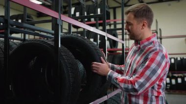 Ekose gömlekli bir adam, bir oto yedek parça dükkanındaki bir mağazada lastiği inceler, çeşitli lastik seçenekleriyle dolu raflarla çevrilidir..