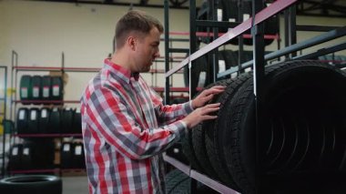 Ekose tişörtlü bir adam mağazadan bir kış lastiği alır araba yedek parça dükkanında lastiklerle çevrili bir satıcıyı arar..