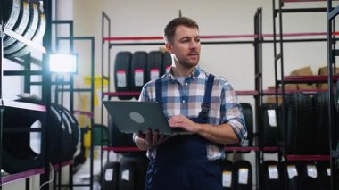 Tulumlu bir erkek işçi lastik dükkanındaki envanterleri kontrol etmek için dizüstü bilgisayar kullanıyor. Hisse senetlerini yönetiyor, ürünleri düzenliyor ve ulaşılabilirliği sağlıyor..