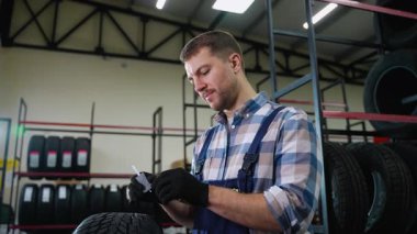 Tulumlu ve eldivenli bir lastik dükkânı çalışanı ölçü aleti kullanarak lastik derinliğini kontrol eder. Depodaki lastiklerin kalitesini ve güvenliğini sağlıyor..
