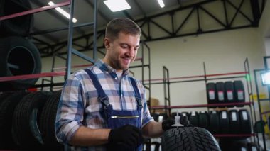 Tulum ve eldiven giyen neşeli bir lastik dükkânı işçisi bir gösterge kullanarak derinlik ölçer, stoktaki lastiklerin güvenliğini ve kalitesini sağlar..