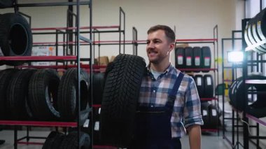 Tulum giymiş neşeli bir işçi, lastik dolu raflarla çevrili bir otomotiv mağazasında ya da depoda omzunda bir lastik tutar..