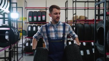 Ekose gömlekli ve tulumlu bir lastik dükkanı çalışanı, bir otomotiv mağazasında iki lastik taşır ve onları müşteri alımı ya da montaj için hazırlar..