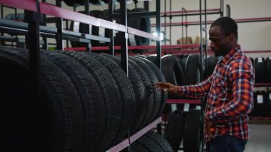 Ekose gömlekli bir adam bir otomotiv mağazasında sergilenen lastikleri dikkatle inceliyor. Aracı için en iyi seçeneği göz önünde bulundurarak seçenekleri değerlendiriyor..