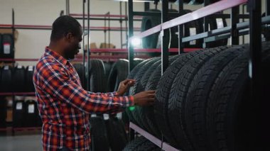 Ekose gömlekli Afro-Amerikalı bir adam bir mağaza rafındaki lastiği inceliyor. Aracı için en iyi seçeneği dikkatlice seçiyor..