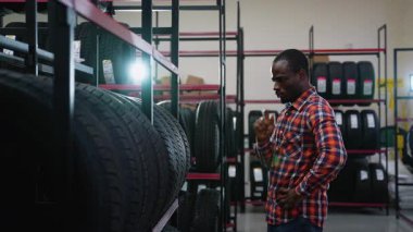 Ekose gömlekli Afro-Amerikalı bir adam otomobil yedek parça dükkanından lastikleri dikkatle seçer, aracının seçeneklerini karşılaştırır.