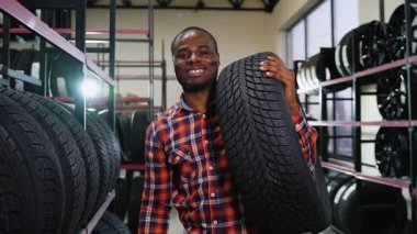 Ekose gömlekli, gülümseyen bir Afrikalı Amerikalı, araba lastikleri ve aksesuarlarla dolu raflarla çevrili bir oto tamirhanesinde lastik taşıyor..