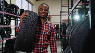 Ekose gömlekli, gülümseyen bir Afrikalı Amerikalı, araba lastikleri ve aksesuarlarla dolu raflarla çevrili bir oto tamirhanesinde lastik taşıyor..