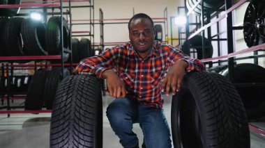 Ekose gömlekli gülümseyen bir adam bir oto yedek parça mağazasında iki yeni lastik arasına çömelmiş stoklanmış raflar arasındaki alımından memnun görünüyor..