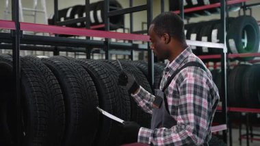 Eldivenli ve üniformalı bir depo çalışanı, otomotiv deposunda bir not panosu ve kalemle envanteri incelerken lastikleri kontrol eder..