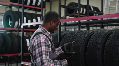 Ekose gömlekli ve eldivenli bir işçi deponun tekerlerini incelerken envanter sayımını da not ve kalem kullanarak yapıyor.