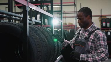 Ekose gömlekli ve tulumlu bir erkek işçi bir otomotiv deposunda elinde bir pano tutarken depodaki lastikleri kontrol ediyor..