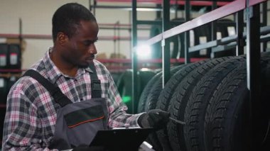 Ekose gömlekli ve tulumlu Afrikalı Amerikalı bir işçi otomotiv mağazasında ya da depoda lastik envanterini kağıt ve kalemle kontrol ediyor..