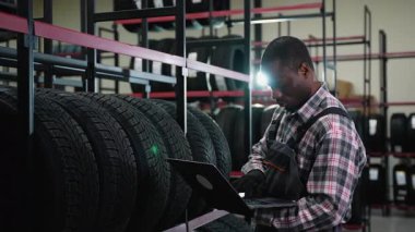 Ekose gömlekli ve tulumlu bir Afrikalı Amerikalı işçi, organize raflarla dolu bir lastik deposunda envanter işletirken dizüstü bilgisayar kullanıyor..