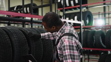İş tulumu ve eldiven giymiş Afrikalı bir Amerikalı bir işçi bir depodaki bir rafta lastik teftişi yapıyor. Kalite ve envanter yönetimi sağlanıyor..