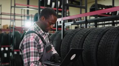 Önlüklü ve eldivenli bir Afro-Amerikalı depo çalışanı dizüstü bilgisayardaki lastik envanterini kontrol ederek hisse senedi doğruluğunu ve envanter yönetimini garanti altına alıyor..