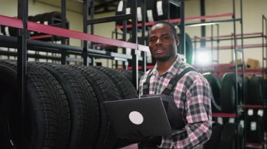 İş tulumu ve eldiven giymiş Afrikalı bir depo çalışanı bir otomotiv mağazasında verimli bir hisse yönetimi sağlamak için lastik stoklarını kontrol etmek için dizüstü bilgisayar kullanıyor..