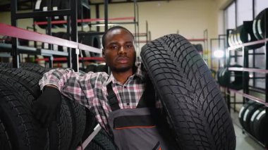 Ekose gömlekli ve tulumlu bir Afrikalı Amerikalı işçi bir otomotiv dükkanında lastik tutuyor. Etrafı satılık çeşitli lastiklerle dolu..