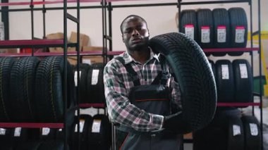 Tulum giymiş neşeli bir işçi, lastik dolu raflarla çevrili bir otomotiv mağazasında ya da depoda omzunda bir lastik tutar..