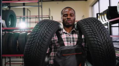 Ekose gömlekli ve tulumlu Afro-Amerikan bir işçi bir otomotiv mağazasında iki lastik bulundurur ve onları müşteri alımına hazırlar..